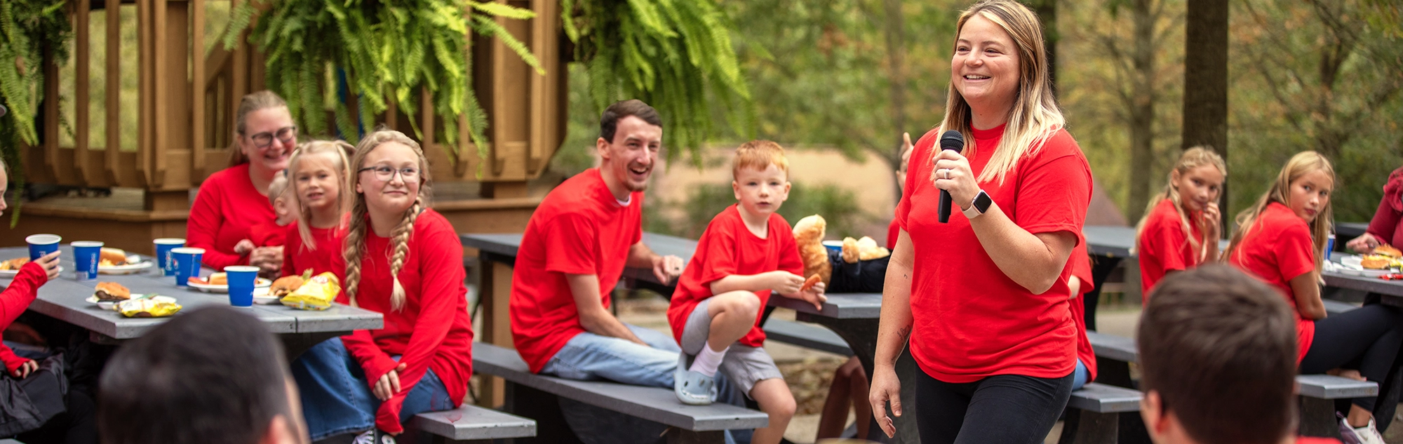 Guests in red shirts sit at picnic tables listening to a woman speaking to them.
